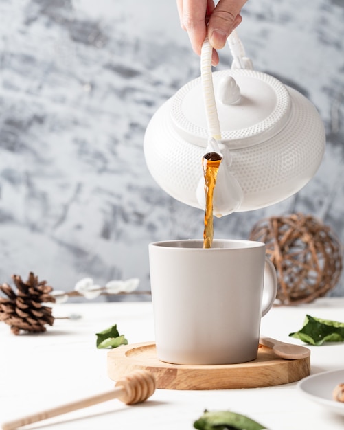 Vertikaler Schuss eines Tees, der von einem Kessel zu einer weißen Tasse mit einem Holzlöffel fließt