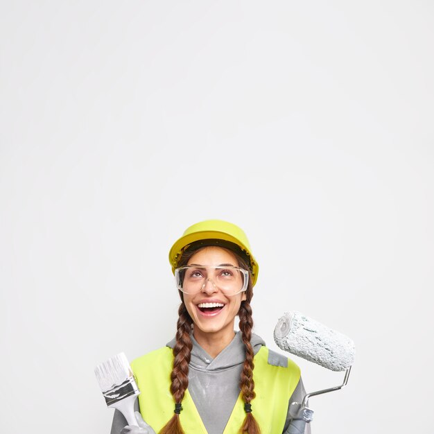 Vertikales Bild einer positiven Bauarbeiterin hält Ausrüstung zum Bemalen von Wänden in Arbeitskleidung, die sich oben mit fröhlichem Ausdruck isoliert über weißer Wand mit leerem Raum konzentriert