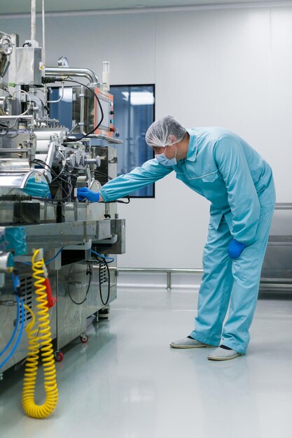 Vertikales Bild eines gebogenen kaukasischen Wissenschaftlers im blauen Laboranzug, der versucht, eine Fertigungsmaschine mit Wellen in einer sauberen Fabrik zu reparieren