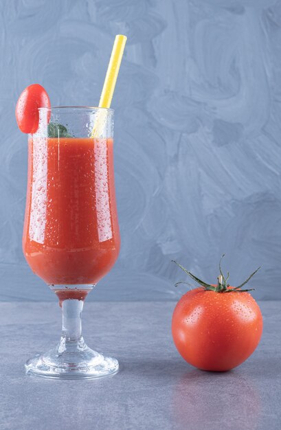 Vertikales Foto des Glases des frischen Tomatensaftes und der Tomate auf einem grauen Hintergrund.