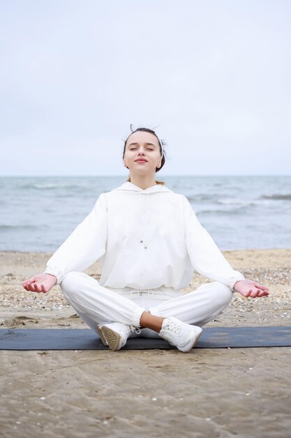 Vertikales Foto einer jungen Dame, die auf einer Matte sitzt und am Strand meditiert Foto in hoher Qualität