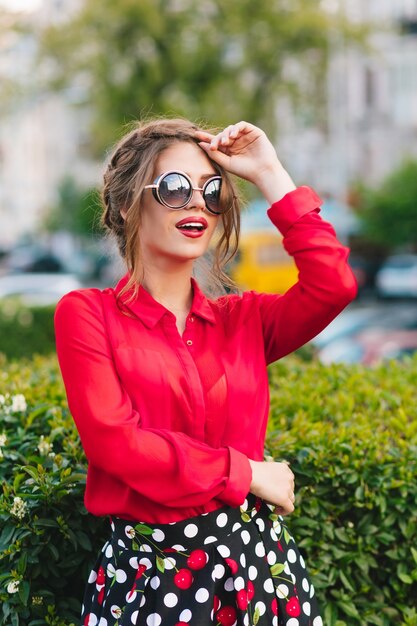 Vertikales Porträt des hübschen Mädchens in der Sonnenbrille, die zur Kamera im Park aufwirft. Sie trägt eine rote Bluse, einen schwarzen Rock und eine schöne Frisur. Sie schaut weit weg.