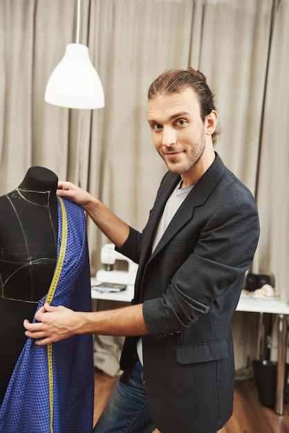 Vertikales Porträt des jungen attraktiven professionellen kaukasischen männlichen Designers im stilvollen Outfit, das an der neuen Kleiderkollektion für folgende Modenschau arbeitet und Ausschnittgröße mit Maßband prüft