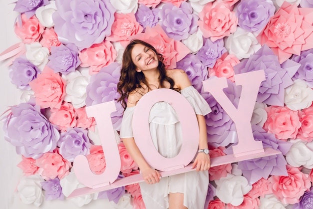 Vertikales Porträt des niedlichen brünetten Mädchens. Sie steht und hält das Holzwort JOY und lächelt breit. Sie hat rosa Hintergrund mit Blumen bedeckt