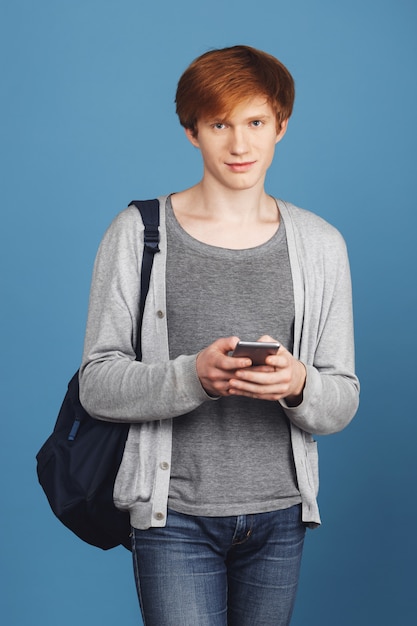 Vertikales Porträt des schönen ernsten jungen Studenten Kerl mit roten Haaren im lässigen Outfit mit schwarzem Rucksack, der mit Freundin per Telefon, mit ruhigem Ausdruck plaudert