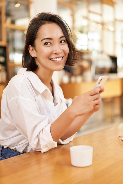 Vertikales Porträt einer stilvollen asiatischen Frau, die im Café sitzt, Kaffee trinkt und Smartphone benutzt