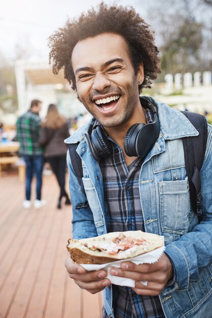 Vertikales Porträt eines charmanten, nicht rasierten, dunkelhäutigen Mannes, der ein leckeres Sandwich hält, während er mit einem Rucksack im Park spazieren geht oder an einem Lebensmittelfest teilnimmt, laut lacht und gute Laune ausdrückt