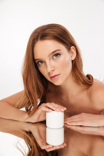 Kostenloses Foto vertikales schönheitsporträt der ingwerfrau mit dem langen haar stützt sich auf spiegeltabelle beim halten der flasche körpercreme und schauen