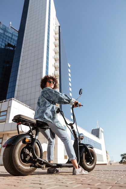 Vertikales Seitenansichtbild der sorglosen lockigen Frau in der Sonnenbrille, die auf dem modernen Motorrad draußen sitzt und weg schaut