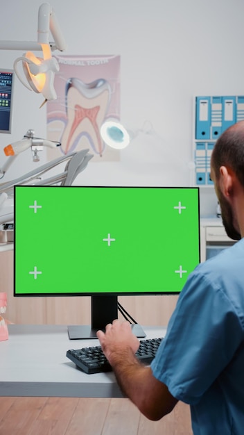 Vertikales Video: Assistent, der in der Zahnarztpraxis einen horizontalen grünen Bildschirm auf dem Computer betrachtet. Mann, der als Zahnpflegespezialist mit Mockup-Hintergrund und isolierter Vorlage auf dem Monitor arbeitet