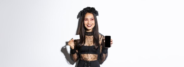 Verträumt lächelndes Mädchen, das wegschaut, während es nachdenkt, Kreditkarte und Handy mit Hallowe zu zeigen