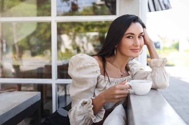 Verträumte attraktive Frau in modischem Outfit, die eine Tasse Kaffee hält und die Kamera genießt, die die perfekte sonnige Morgenfrühstückszeit genießt. Sexy Frau im Urlaub trinkt Cappuccino im Hotelrestaurant