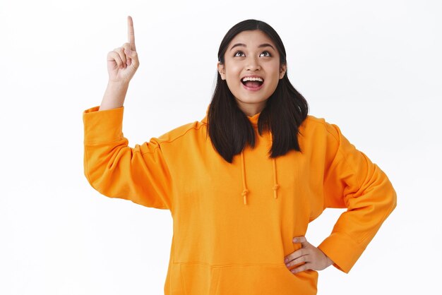 Verträumte, aufgeregte, lächelnde asiatische Frau in orangefarbenem Kapuzenpulli beobachtet etwas Cooles, das nach oben schaut und auf die obere Werbung nach oben zeigt, Promo-Werbung, die auf weißem Hintergrund steht