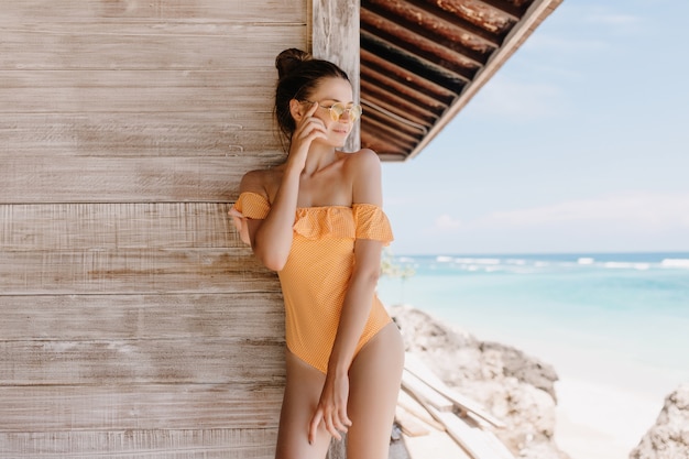 Verträumte braunhaarige Frau in der Sonnenbrille, die im Resort ruht und Blick auf den Himmel genießt. Außenaufnahme des reizenden schlanken Mädchens in der Badebekleidung, die nahe Holzwand mit glücklichem Gesichtsausdruck aufwirft.