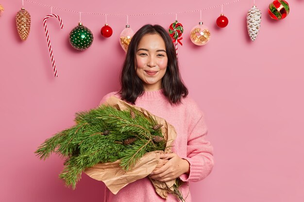 Verträumte erfreute asiatische Frau hält handgemachten Neujahrsstrauß aus grünen Fichtenzweigen gekleidet in lässigem Pullover bereitet sich auf Winterferien vor