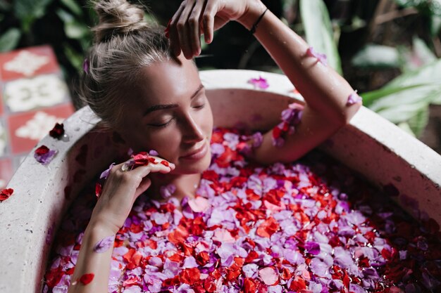 Verträumte Frau, die mit geschlossenen Augen im Bad voller rosa Rosen liegt. Überkopfaufnahme der romantischen Dame mit gebräunter Haut, die während des Spa am Morgen abkühlt.