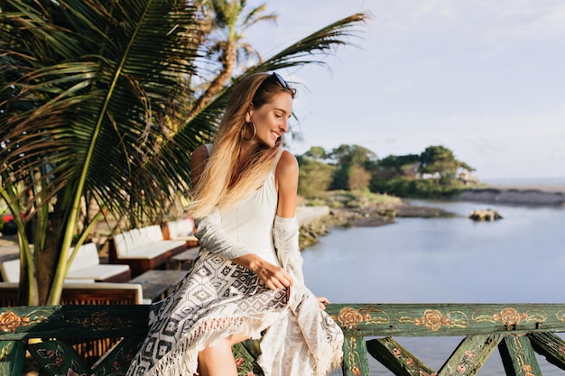 Verträumte junge Frau, die in der Nähe eines exotischen Baums auf Meereshintergrund sitzt Outdoor-Foto einer angenehmen Dame in weißer Kleidung, die die Aussicht auf die Natur im Resort genießt