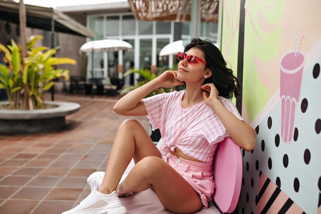 Verträumte junge Frau in Turnschuhen, die im Café im Freien sitzt Gebräuntes Mädchen mit Tätowierung, das an warmen Tagen auf der Straße posiert