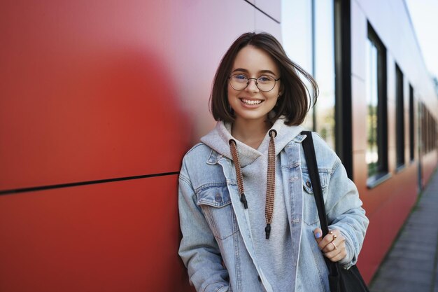 Verträumte süße lächelnde Frau mit Brille tauscht Studentin aus, die auf dem Campus spazieren geht und eine Tragetasche hält, die mit der Kamera sorglos und fröhlich aussieht, Konzept der Jugend, moderne Generation und Karriere