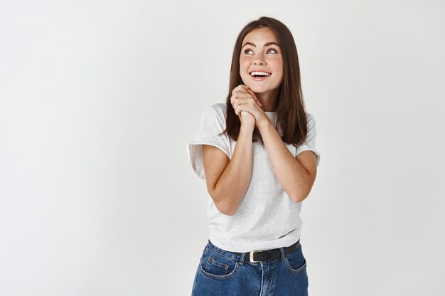 Verträumte und leidenschaftliche Frau, die mit Bewunderung in die obere linke Ecke schaut, sich etwas vorstellt und lächelt, über weißer Wand stehend