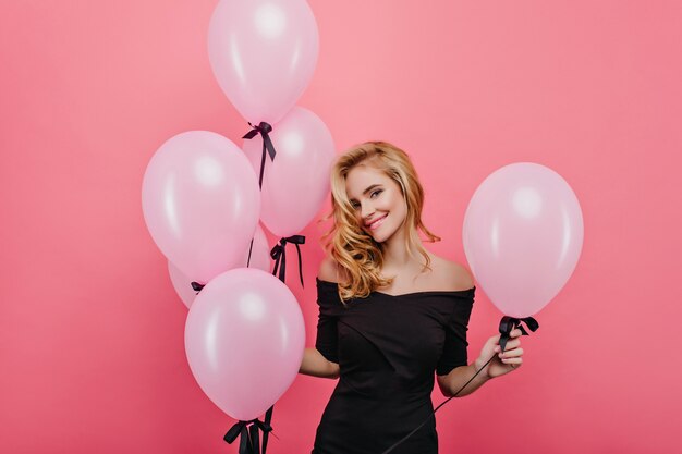 Verträumte weiße Frau mit dem niedlichen Gesicht, das auf rosa Wand mit Luftballons aufwirft. Elegantes blondes weibliches Modell, das Geburtstag mit Partei feiert.