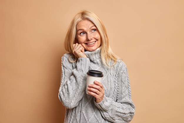 Verträumte zerknitterte Rentnerin mit blondem Haar, minimalem Make-up in warmem grauem Pullover, träumt von etwas Angenehmem und trinkt Kaffee.
