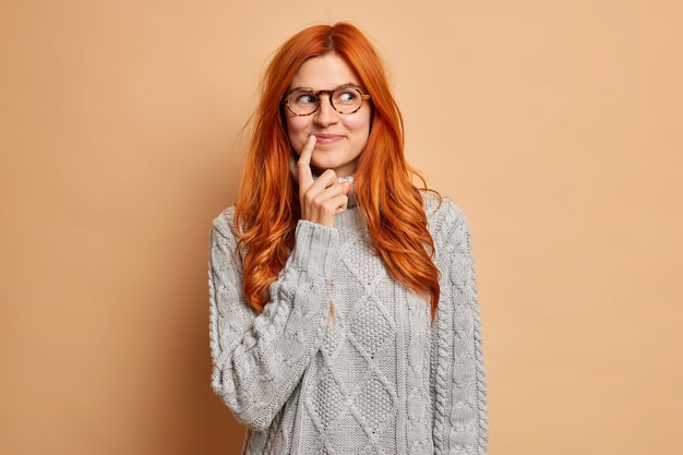 Kostenloses Foto verträumte zufriedene rothaarige frau hält zeigefinger in der nähe von lippenwinkeln konzentriert beiseite trägt nachdenklich grauen strickpullover.