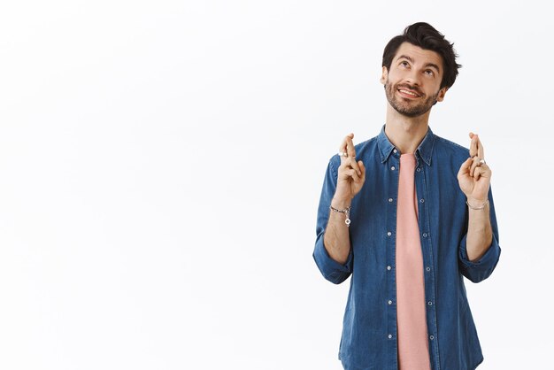 Verträumter, charmanter bärtiger 25er-Typ im Hipster-Shirt-T-Shirt, der Tagträumen oder fantasierende Kreuzfinger für viel Glück aufblickt und Wünsche lächelnd nachdenklicher Imaging-Traum wahr werden lässt, weißer Hintergrund