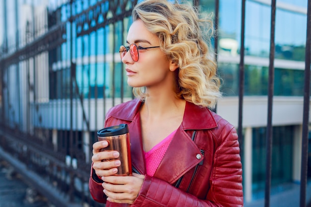 Verträumtes blondes Mädchen, das die Straßen geht, Kaffee oder Cappuccino trinkend. Stilvolles Herbstoutfit, Lederjacke und Strickpullover. Rosa Sonnenbrille.