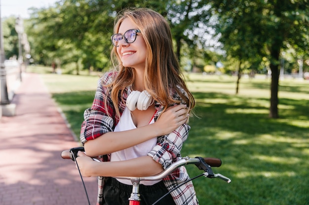 Verträumtes kaukasisches Mädchen mit dem Fahrrad, das mit Lächeln herumschaut. Hübsche junge Dame in den Gläsern, die im Park stehen.