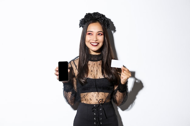 Verträumtes lächelndes Mädchen, das weg schaut, während denkend, Kreditkarte und Handy zeigend, Halloween-Gothic-Kleid tragend.