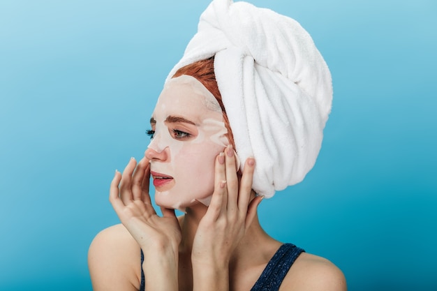 Verträumtes Mädchen, das Gesichtsmaske lokalisiert auf blauem Hintergrund anwendet. Studioaufnahme der jungen Dame mit Handtuch auf dem Kopf weg schauend.