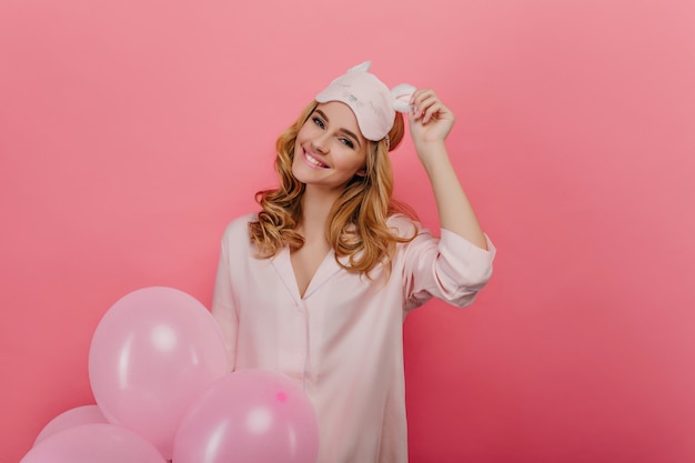Verträumtes Mädchen, das ihre Augenmaske mit Lächeln berührt, während es auf rosa Wand aufwirft. lachende erstaunliche Frau im Pyjama, Partyballons haltend.