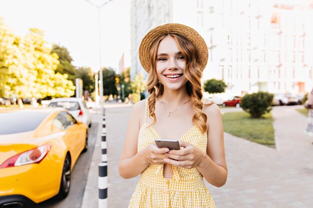 Verträumtes Mädchen im gelben Weinlesekleid, das positive Gefühle während des Gehens ausdrückt. Erstaunliche Frau mit welligem Haar, das Smartphone hält.