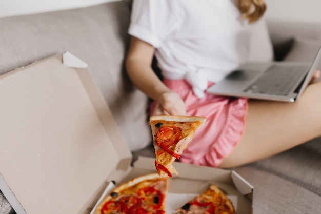 Kostenloses Foto verträumtes mädchen in den rosa shorts, die auf sofa sitzen und ein stück pizza halten. innenaufnahme der raffinierten weißen frau, die mit computer isst und arbeitet.