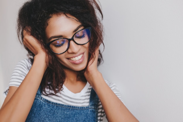 Verträumtes mädchen in der stilvollen brille, die mit geschlossenen augen und erstaunlichem lächeln auf weißer wand aufwirft