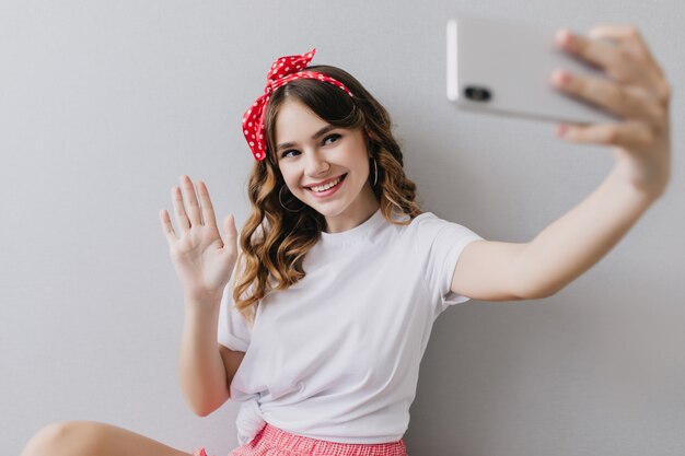 Verträumtes Mädchen mit gewellter Frisur, die mit Lächeln aufwirft. Innenaufnahme der wundervollen jungen Dame im weißen lässigen T-Shirt, das Selfie macht.