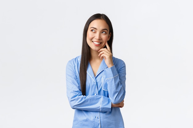 Verträumtes nachdenkliches asiatisches Mädchen in blauen Pyjamas mit interessanter Idee, Blick in die obere linke Ecke auf Kommentarblase, Lächeln erfreut als Denken, Tagträumen vor dem Schlafengehen, weißer Hintergrund.