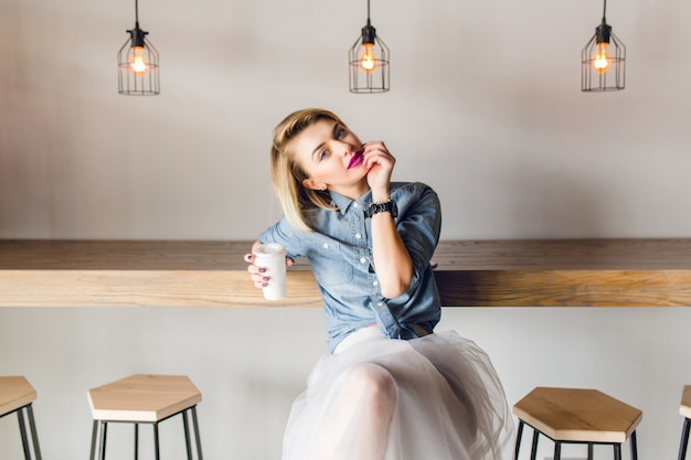 Verträumtes stilvolles Mädchen mit blonden Haaren und rosa Lippen, die in einem Café mit Holzstühlen und Tisch sitzen. Sie hält eine Tasse Kaffee