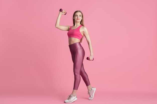 Vertrauen. Schöne junge Sportlerin üben, einfarbiges rosa Porträt. Kaukasisches Modell der sportlichen Passform mit Gewichten. Bodybuilding, gesunder Lebensstil, Schönheits- und Aktionskonzept.