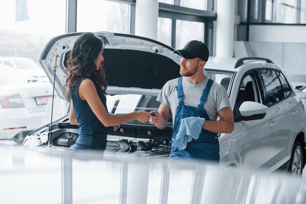 Vertrauen und Beruf. Frau im Autosalon mit dem Angestellten in der blauen Uniform, die ihr repariertes Auto zurücknimmt