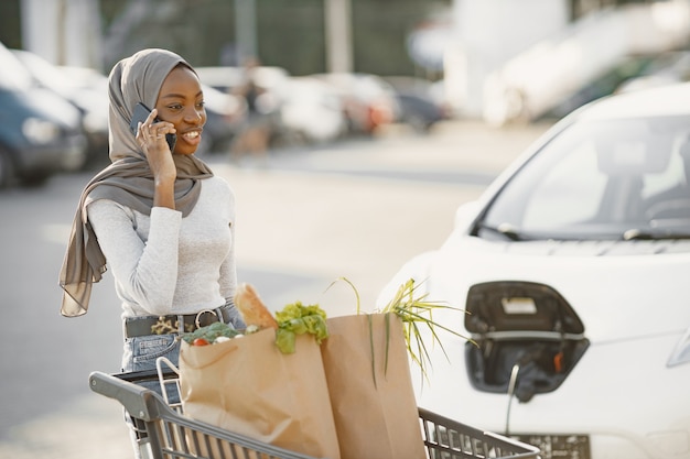 Verwenden des Smartphones während des Wartens. Afrikanische ethnische Frau auf der Ladestation für Elektroautos tagsüber. Nagelneues Fahrzeug.