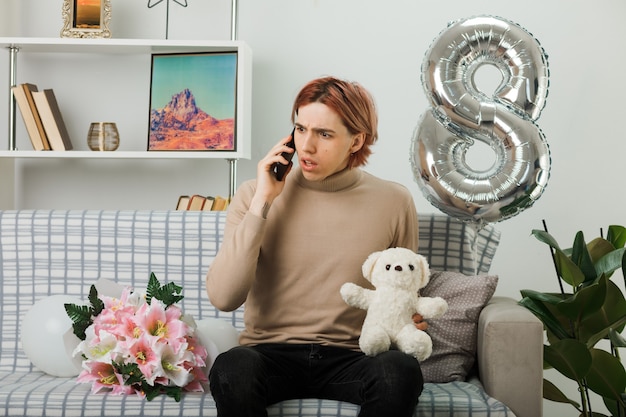 Verwirrt aussehender, gutaussehender kerl am glücklichen frauentag, der teddybären hält, spricht am telefon, das auf dem sofa im wohnzimmer sitzt
