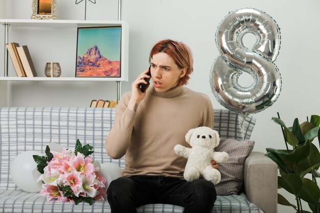 Verwirrt aussehender, gutaussehender Kerl am glücklichen Frauentag, der Teddybären hält, spricht am Telefon, das auf dem Sofa im Wohnzimmer sitzt