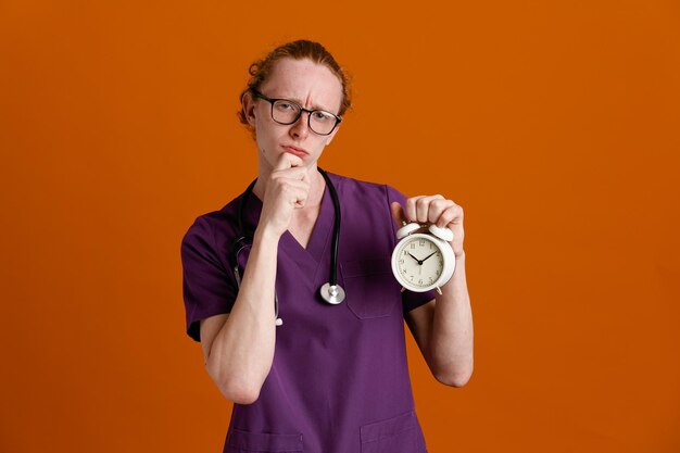 verwirrt packte Kinn hält Wecker junger männlicher Arzt in Uniform mit Stethoskop isoliert auf orangefarbenem Hintergrund