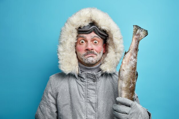 Verwirrt überrascht gefrorener Mann verbringt lange Stunden im Freien während eines strengen kalten Tages gekleidet in graue Winterjacke und Handschuhe hält Fisch trägt Skibrille.