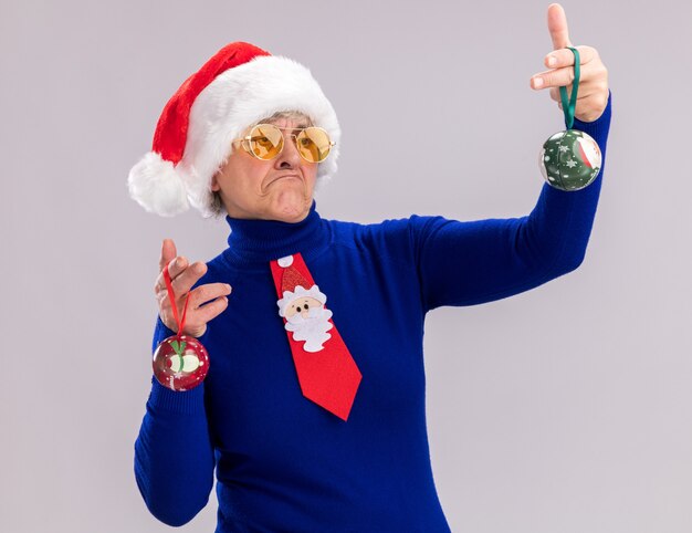 Verwirrte ältere Frau in Sonnenbrille mit Weihnachtsmütze und Weihnachtsmann-Krawatte, die Glaskugelverzierungen einzeln auf weißer Wand mit Kopienraum hält und betrachtet