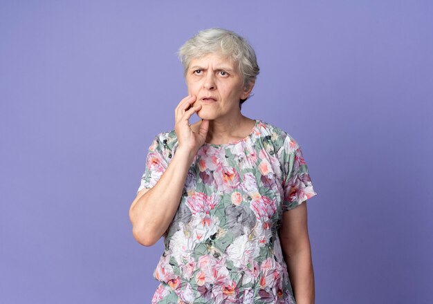 Verwirrte ältere Frau legt Hand auf Kinn isoliert auf lila Wand