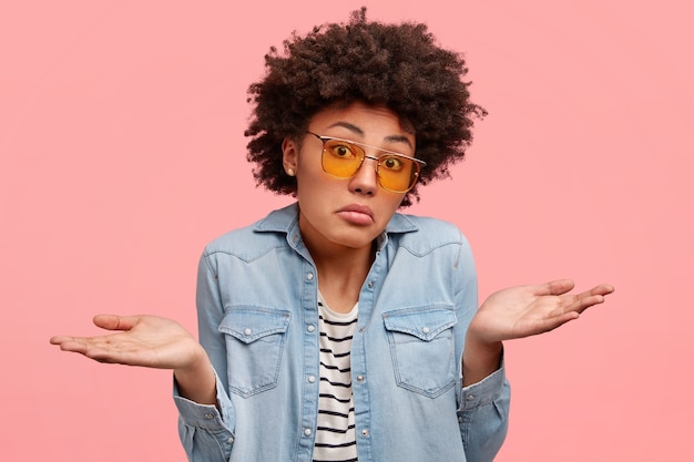 Kostenloses Foto verwirrte afroamerikanerin mit ahnungslosem gesichtsausdruck, zuckt mit den schultern, weiß keine antwort, sieht keinen ausweg, trägt modische jeanskleidung und sonnenbrille, modelle drinnen über rosa wand