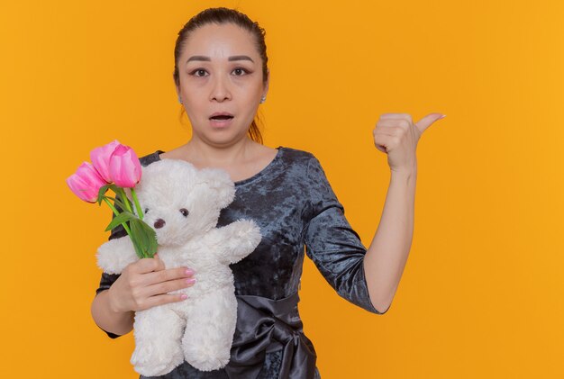 Verwirrte asiatische Frau, die Strauß der rosa Tulpen und des Teddybären hält, der vorne zeigt, zeigt zurück mit Daumen, der internationalen Frauentag feiert über orange Wand